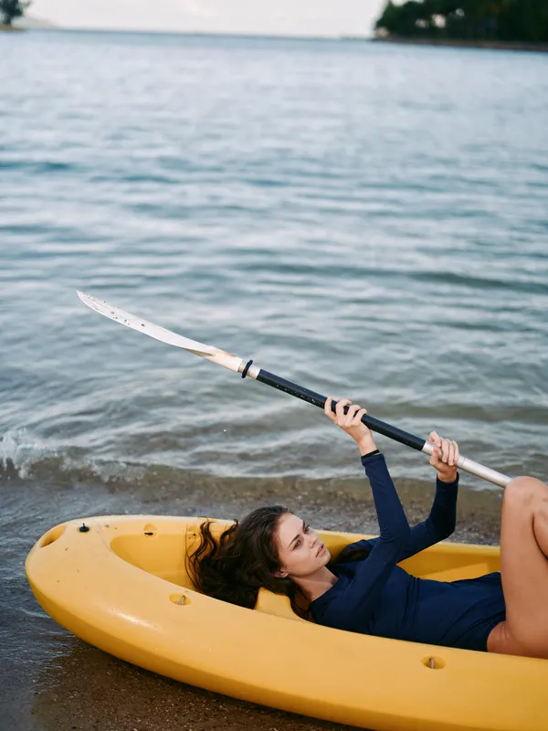 Young woman lying in yellow kayak on the shore holding paddle, relaxing near calm water shore on a warm day.