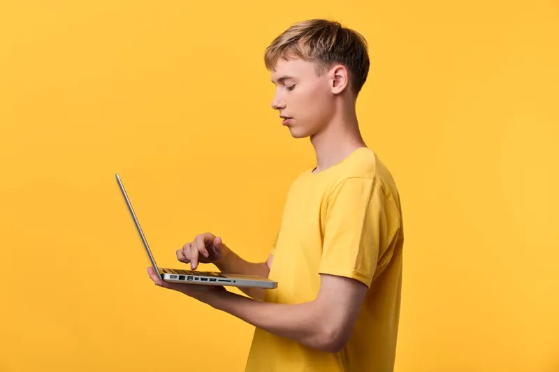 Young man in a yellow shirt using a laptop for typing and digital work, showcasing modern technology and focused concentration against a bright yellow backdrop.