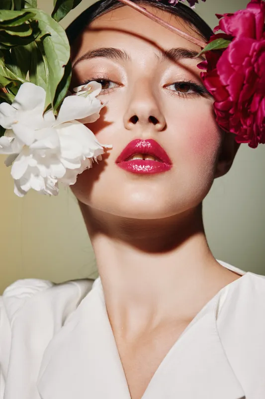 Teen woman with glossy red lips surrounded by colorful peony flowers. Soft natural light highlights radiant skin and shiny makeup creating fresh beauty and fashion concept.