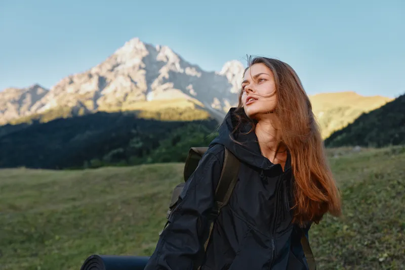 A solo hiker woman stands outdoors in a mountainous landscape. She carries a backpack and gazes toward the distant peaks with calm confidence and natural serenity.