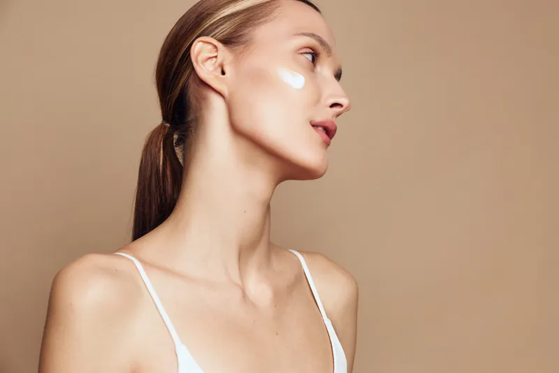 Female portrait in side profile with cream on cheek, neutral beige backdrop, soft lighting, clean skin, emphasizes skincare routine and natural beauty in a studio setting.