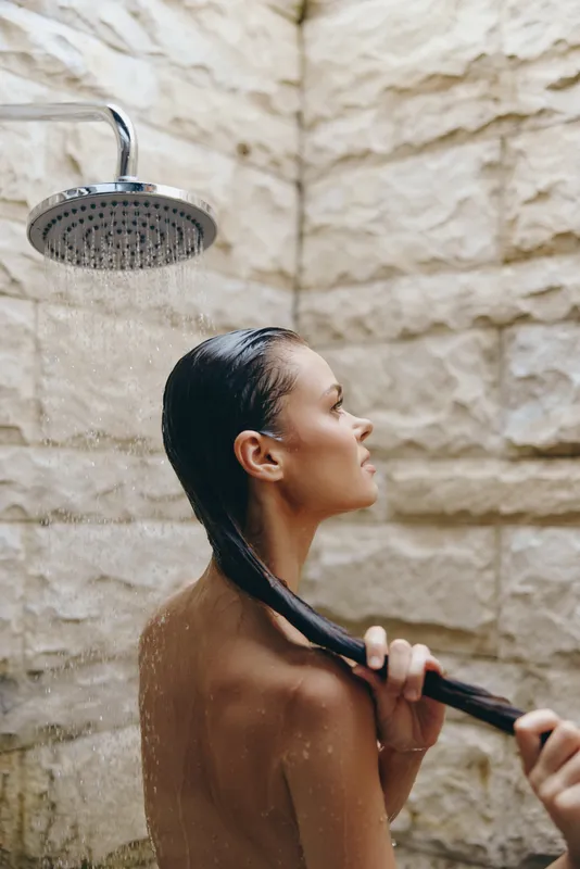 Woman showering under rainfall showerhead with stone wall background, wet hair, and relaxed expression in natural light bathroom setting.