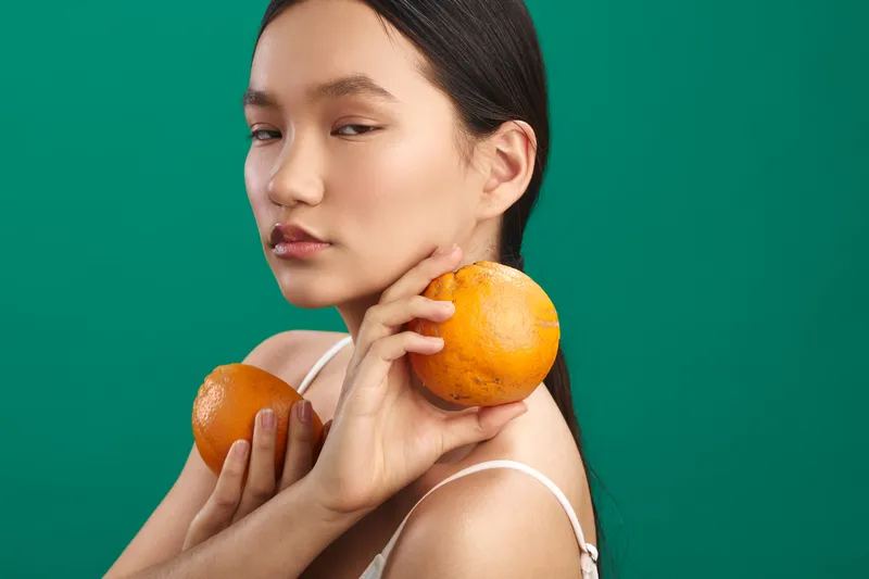 asian woman holds oranges against a green backdrop with soft lighting, conveying calm, wellness, and a fresh lifestyle moment. the vibrant citrus tones complement a minimal studio setting.