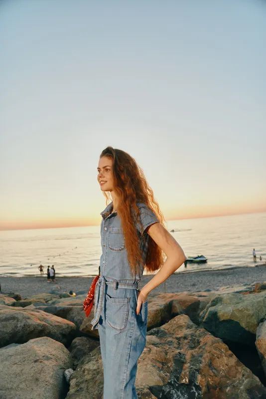 lifestyle woman in modern boho-western denim street style stands on rocky beach at sunset with film color effect, natural light, long curly hair, casual outfit, relaxed mood