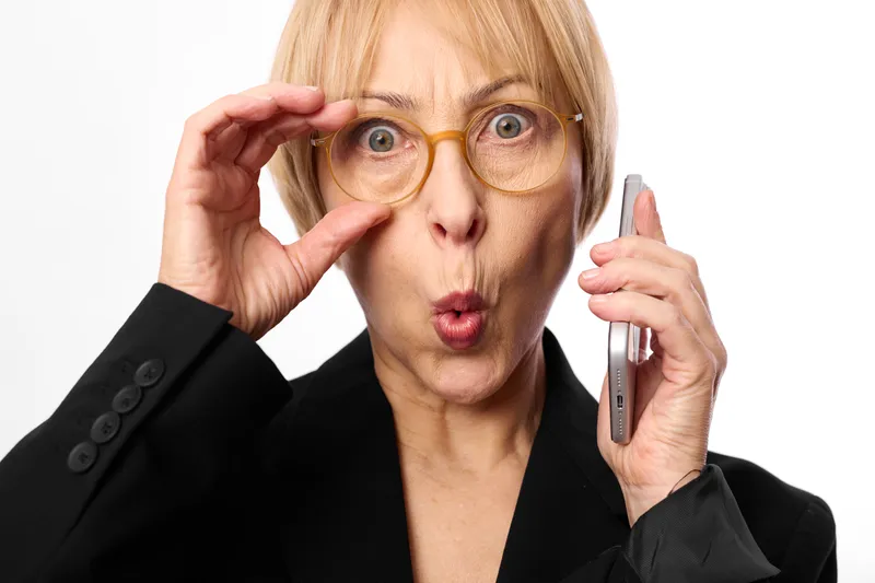 Surprised blonde woman adjusting glasses and holding a smartphone, close up portrait on white background. Business style, makeup and cosmetics concept with soft studio lighting.