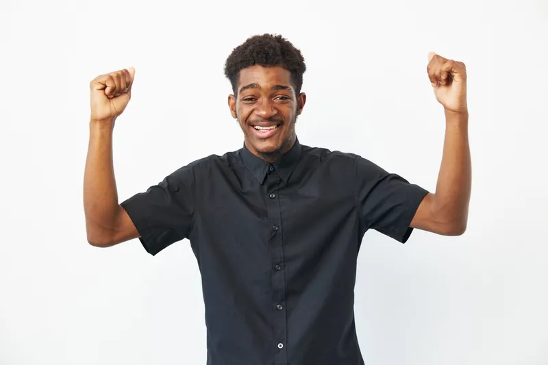 Happy African American man celebrating with raised arms, wearing a casual black shirt, exuding joy and enthusiasm in a bright atmosphere.