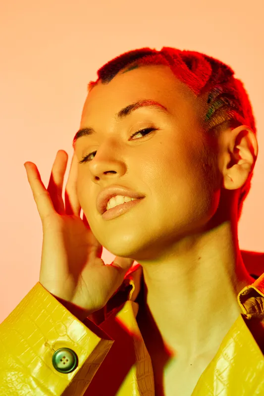 young woman with braided hair posing confidently in yellow textured jacket under warm orange lighting, stylish and vibrant portrait with modern fashion vibe