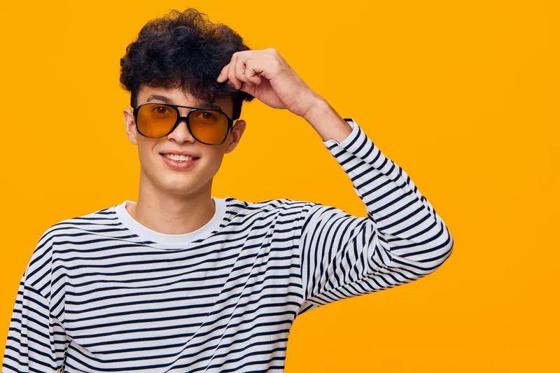 Young man wearing striped shirt and orange sunglasses smiling confidently against bright orange background, casual style and cheerful mood.