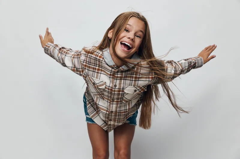 Girl smiling with happy face and long hair wearing plaid shirt and denim shorts posing with arms outstretched against plain background in studio photo.