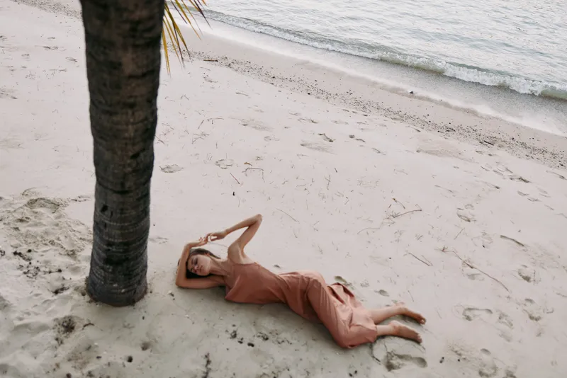 A relaxed figure lies on sunlit sand beside a palm tree, wearing a soft peach dress, gazing upward with a calm, carefree posture in a tranquil seaside setting.