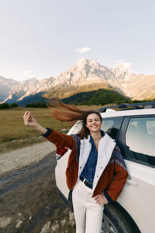 Woman by car in mountains on a roadtrip, smiling and flipping hair beside a white SUV. Casual jacket, scenic landscape and open road evoke adventure, freedom and travel.