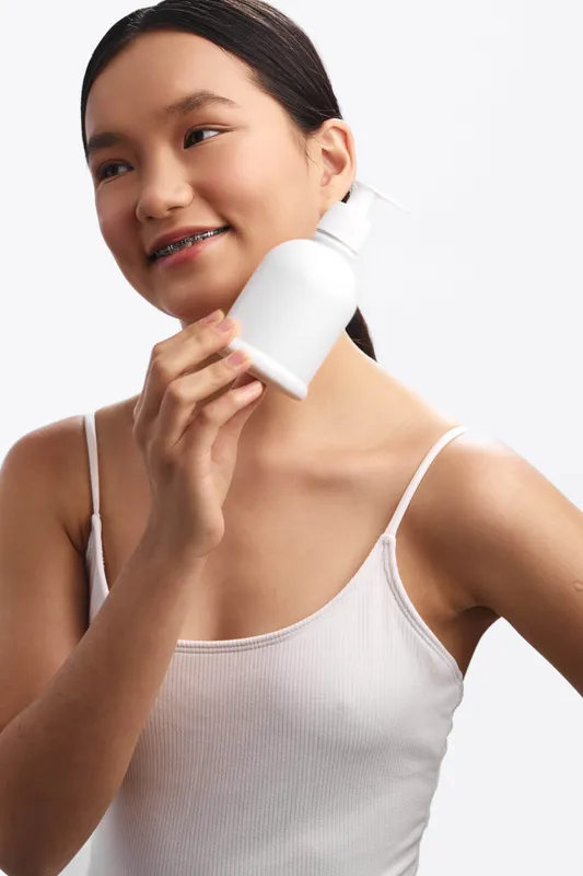 Asian woman smiles gently while holding a white bottle near her face in a clean studio setting. A calm, fresh beauty moment that conveys care, wellness, and everyday self care.