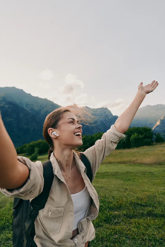 Joyful outdoor woman raising arms in sunlight, hiking vibe with backpack, nature background and mountains, smiling moment, carefree lifestyle meadow field visible