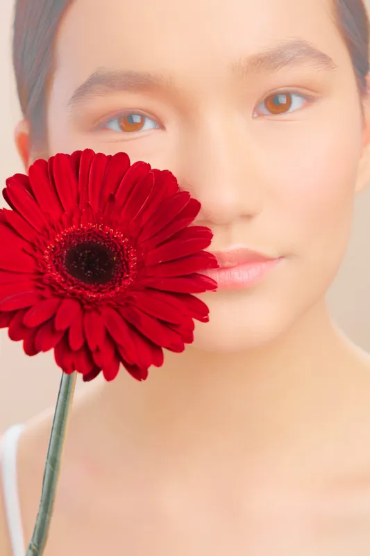 Asian woman gazes softly at the camera, a vivid red flower held near her face, creating an intimate portrait with gentle light and natural color.