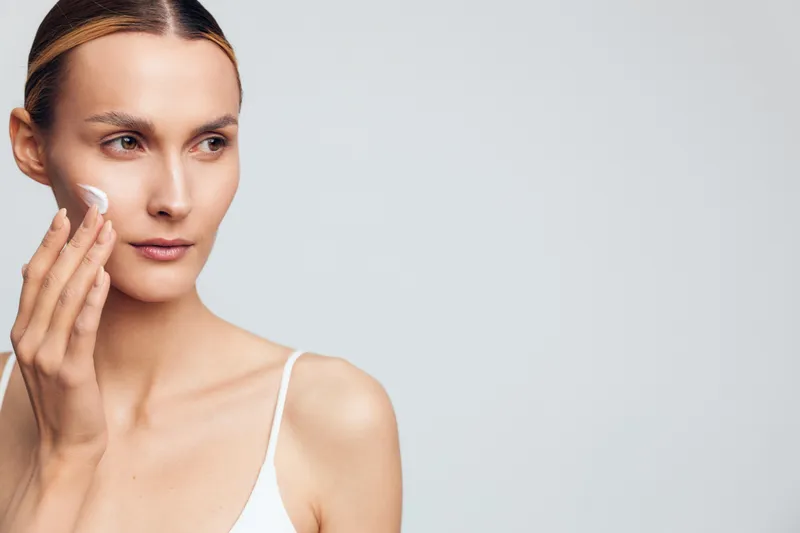 A young woman with clear skin applies white cream to her cheek in a minimalistic setting. The concept focuses on skincare, beauty routine, and natural complexion care.
