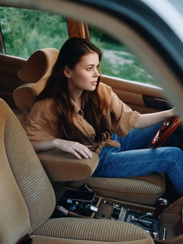 Young woman driving a car with a relaxed expression, wearing a stylish shirt and blue jeans against a natural outdoor backdrop.