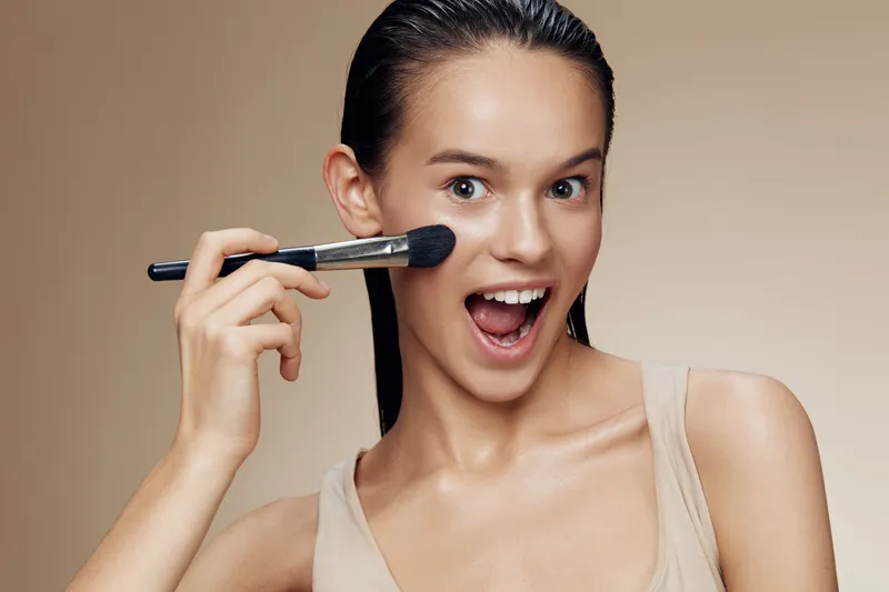 Young cheerful model applying makeup with a brush, showcasing vibrant skin tone and excitement, perfect for beauty and cosmetics concepts