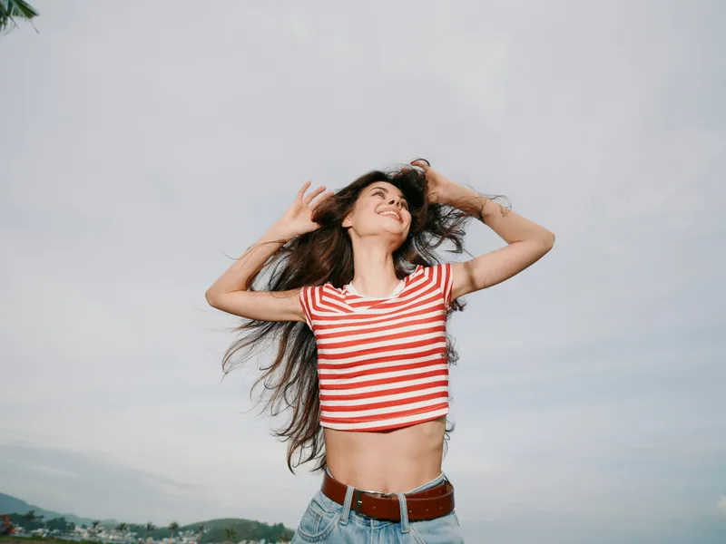 Young woman dances outdoors in a red and white striped crop top and jeans, her hair flowing as she laughs, capturing carefree energy and summer vitality under a clear sky.