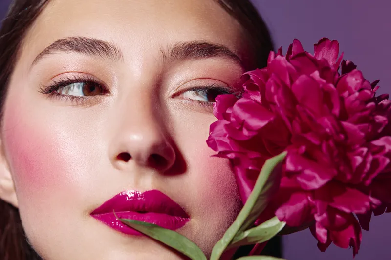 Close-up of woman with bold magenta lipstick and subtle pink eyeshadow holding vibrant pink peony flower. Concept for beauty, creative fashion, and makeup artistry inspiration.