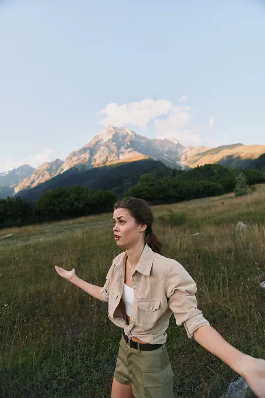 Woman outdoors on a meadow with distant mountains, wearing neutral clothing, arms open in a carefree pose, conveying exploration and freedom and confident moments.