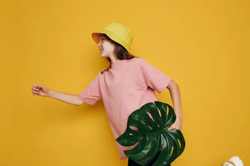 Young woman in a pink t-shirt and yellow bucket hat holding a large green leaf. She looks happy and stands against a solid yellow background. Concept for nature and fashion.