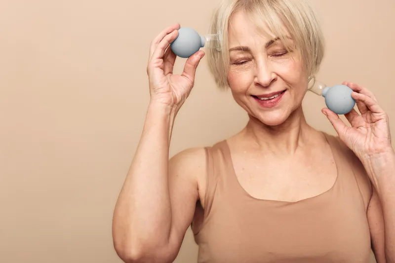 Happy mature woman with closed eyes holds facial massage tools near face, enjoying skincare routine. Beige background and soft lighting create calm and wellness concept for beauty and self-care.