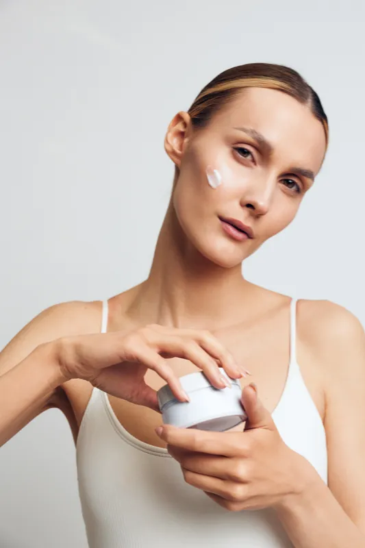 A young woman in a white tank top applies face cream from a jar, showing skincare routine and self care with a neutral background and natural lighting.