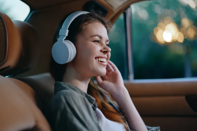 Happy young woman enjoying music with headphones in a car, reflecting joy, relaxation, and solitude in a natural background.
