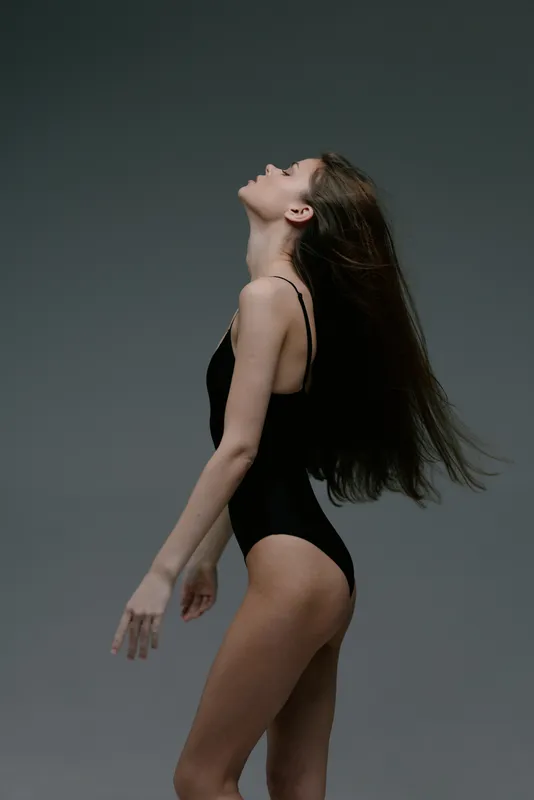 Profile of a slender woman in a black bodysuit leaning back with closed eyes against a dark gray background.