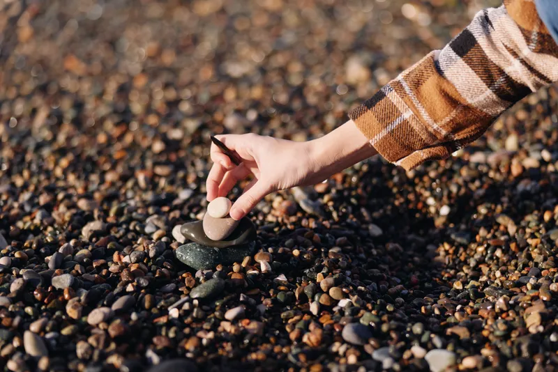 Hand selecting pebbles on a sunlit beach, close up of fingers arranging smooth stones among varied textures and natural colors, a calm outdoor moment of tactile detail and focus