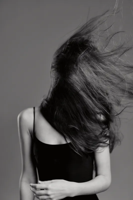 Black and white portrait of a young woman with flowing hair wearing a black dress, captured in motion with artistic style and dynamic energy.
