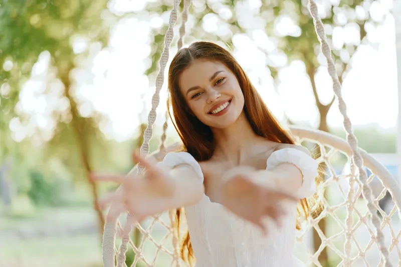 Happy young woman with long hair reaching out from a swing in a bright outdoor setting, wearing a white dress, conveying joy and warmth in a summer atmosphere