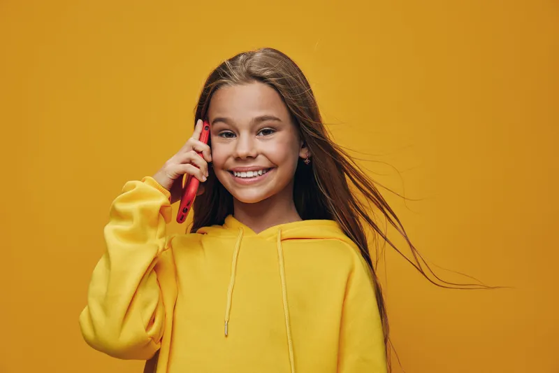 Girl in yellow hoodie talking on smartphone smiling with long hair flowing in wind isolated on yellow background. Child with casual clothing using mobile phone for communication.