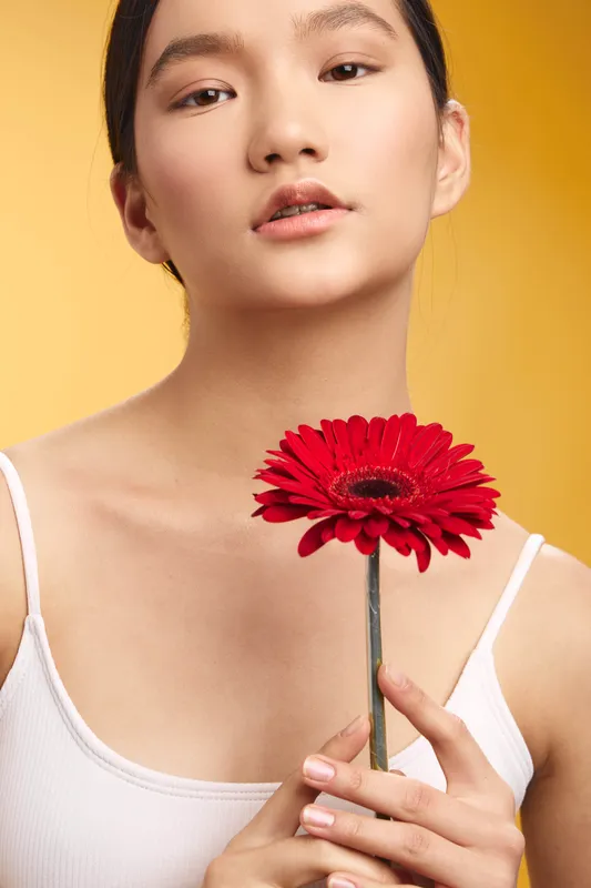 asian woman holds a vibrant red gerbera flower against a warm yellow backdrop, a close up portrait that conveys calm confidence and fresh beauty.