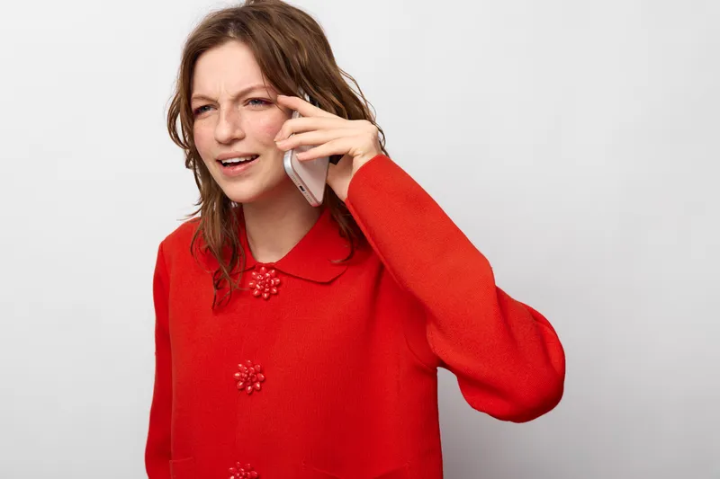 Young woman in red sweater talks on smartphone with confused expression. Isolated on white background. Concept of communication, misunderstanding, and emotional reaction.