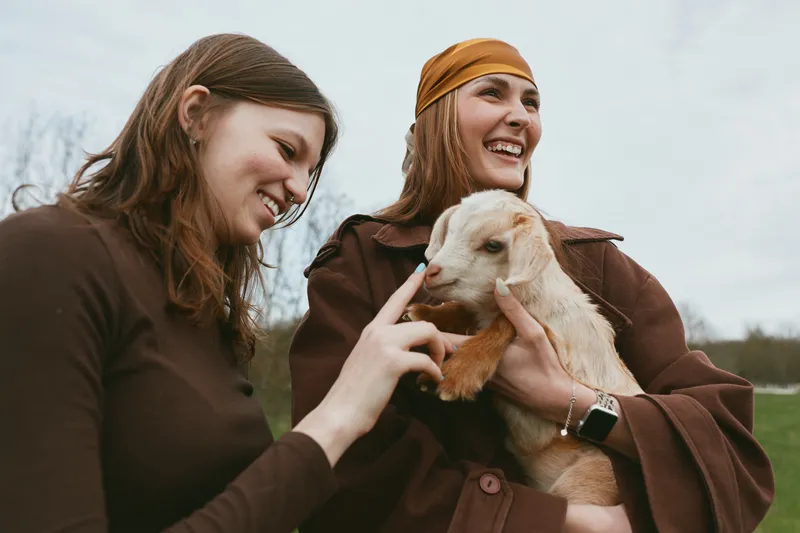 Lifestyle outdoor moment with women and a goat kid, smiling faces, relaxed posture, gentle breeze, natural colors, countryside setting, friendly animals, close bond, lighthearted interaction