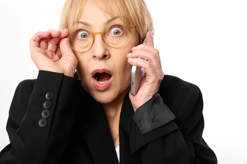 Surprised business woman in black suit holds eyeglasses, gestures during phone communication and call, startled eyes behind glasses, isolated on white studio background.