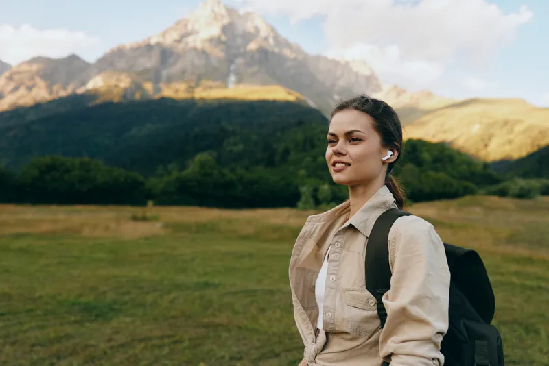 A confident woman hiker with a backpack stands in a sunny mountain landscape, embodying outdoor lifestyle and exploration with open fields and distant peaks behind.