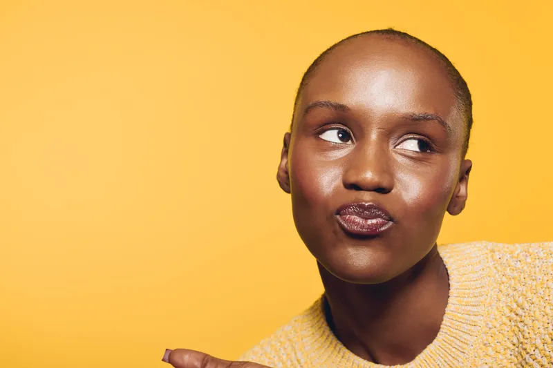 Close-up portrait of a young black woman with dark skin wearing a yellow sweater looking sideways on a vibrant yellow background. Concept of beauty, fashion, and confident expression.