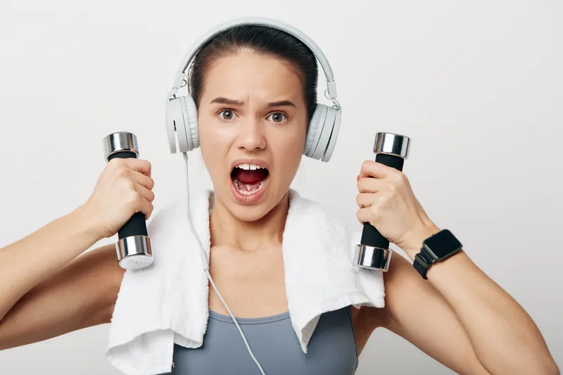 Fitness woman in athletic wear lifts dumbbells with a towel draped over shoulders, headphones on, displaying intensity and motivation during a gym workout session.