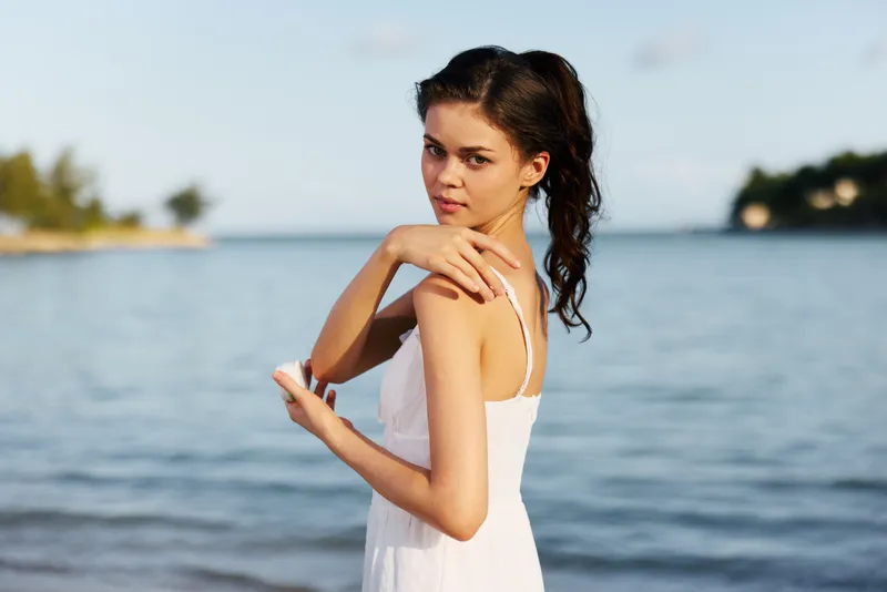 Portrait of a young woman in a white dress by a calm seaside, with soft sunlight, thoughtful pose, loose dark hair, gentle breeze, serene mood, and natural beauty.