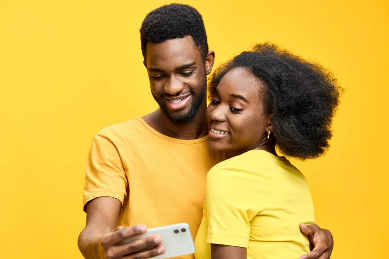 Happy young couple enjoying shared moments while using a smartphone against a vibrant yellow background, showcasing love and connection