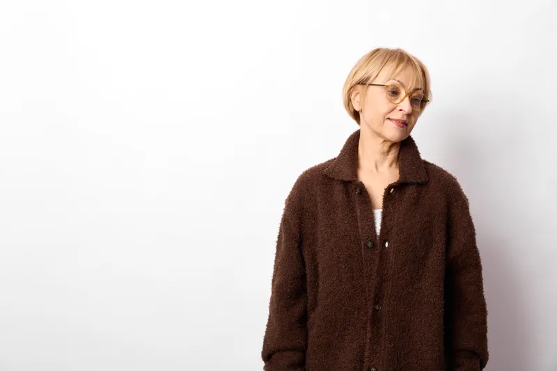 Woman senior smiling portrait in studio, brown coat on white background, relaxed posture, soft light, head and shoulders, calm mood, minimalist composition