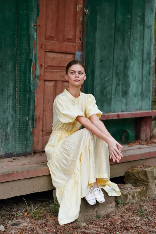 Woman wearing a pale yellow dress sits on stone steps outside a weathered doorway, calm expression and soft natural light creating a serene portrait with rustic charm.