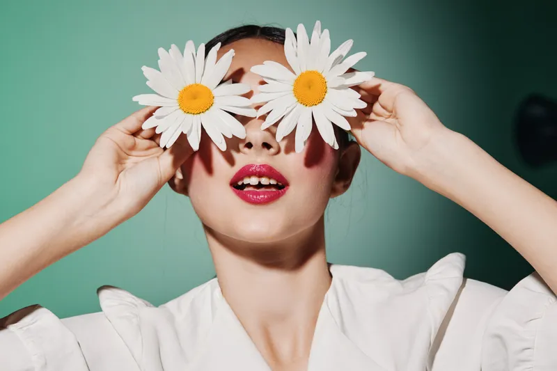 Young woman with bright makeup and red lipstick holds two large flowers over her eyes in a creative fashion portrait. Soft studio lighting and pastel background enhance beauty concept.