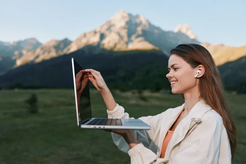 A cheerful woman works outdoors with a laptop in a scenic field beneath mountain peaks. She smiles while typing, conveying focus, productivity, and a refreshing outdoor work vibe.