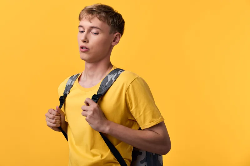 Young student with a backpack wearing a yellow shirt stands before a bright yellow backdrop, conveying casual study vibes, daily routine, learning energy, and readiness for classes.