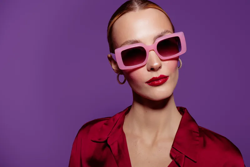 Stylish woman with pink sunglasses and red lipstick poses against purple background. Fashion portrait with confident expression and trendy accessories in bright studio lighting.