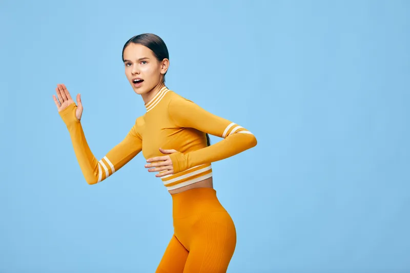 Young athletic woman in bright yellow activewear striking a running pose in studio. Cropped long sleeve top and high waist leggings, dynamic motion, determined expression.