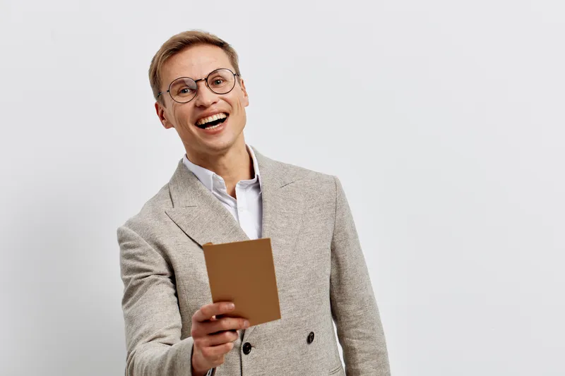 Smiling young man holding book, wearing glasses, beige blazer, white shirt, cheerful expression, studio portrait on plain white background, lifestyle concept